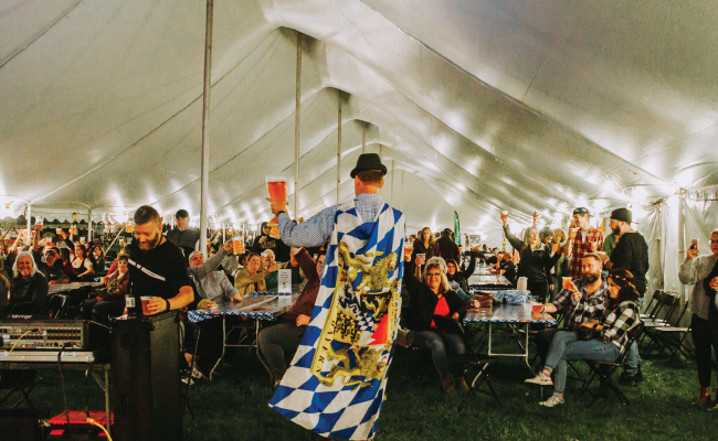 Man walking in a crowd with a German flag on his back, raising a red solo cup under a large tent